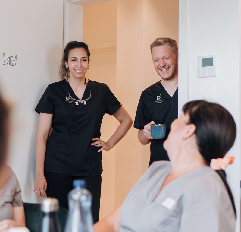 Lisa Kremer (links) und Johannes Well (rechts) besuchen in ausgelassener Stimmung den Pausenraum. Am Tisch sitzen Mitarbeiterinnen.