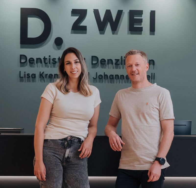 Lisa Kremer (links) und Johannes Well (rechts) stehen lässig vor der Rezeption. Im Hintergrund ist das Logo D.Zwei zu sehen.